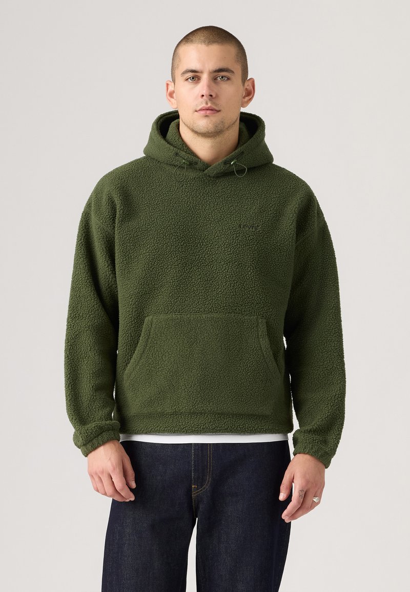 Green fleece hoodie with a textured, soft surface; features a front pocket, adjustable drawstring hood, and ribbed cuffs. Paired with dark jeans.