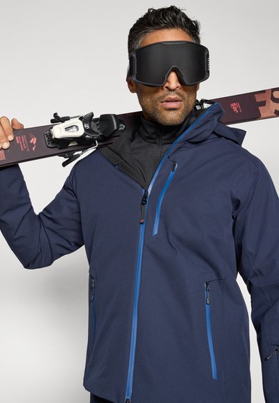 Tamsiai mėlyna slidinėjimo striukė su mėlynomis užtrauktukais, tekstūruota medžiaga, kapišonas ir keli kišenės; modelis laiko slides ir dėvi dideles juodas apsaugines akinius.