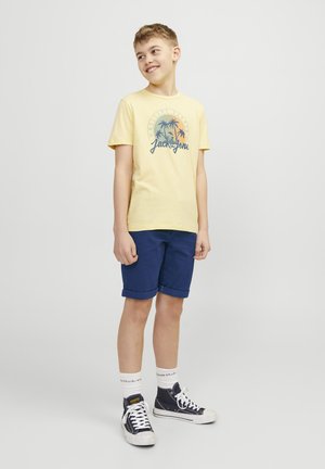 T-shirt jaune avec motif de palmier, accompagnée de shorts navy. Baskets blanches et bleues, et chaussettes blanches avec du texte près du bord.