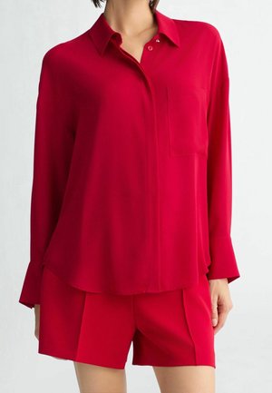 Mujer con blusa roja de botones de manga larga y ajuste holgado, con pantalones cortos rojos a juego, sobre un fondo liso.