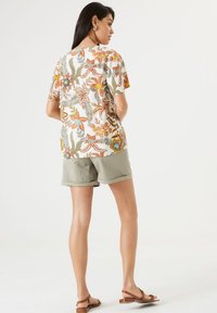 Blusa de manga corta floral en crema, con vibrantes diseños en naranja, azul y verde. Combinada con pantalones cortos en verde oliva y sandalias marrones.