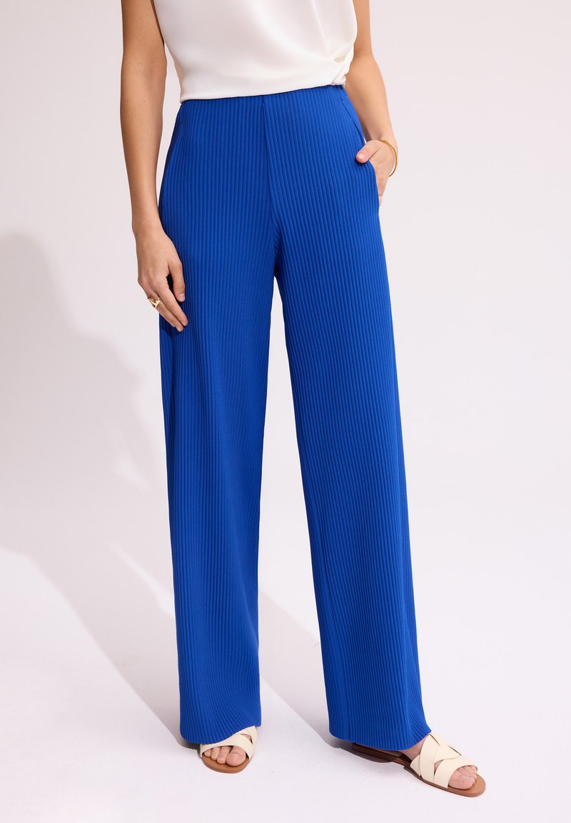 Niebieskie spodnie o szerokich nogawkach z pionowym prążkowanym wzorem, z wysokim stanem i bocznymi kieszeniami, zestawione z beżowymi sandałami na paski.