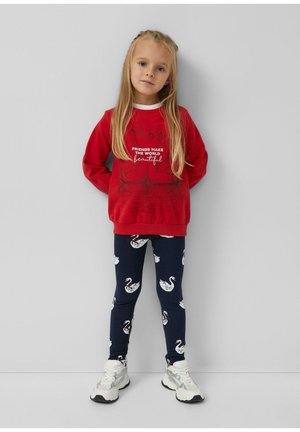 Młoda dziewczyna o długich blond włosach, ubrana w czerwoną bluzę z napisem, granatowe legginsy z białymi nadrukami łabędzi oraz białe trampki, stojąca przy ścianie.