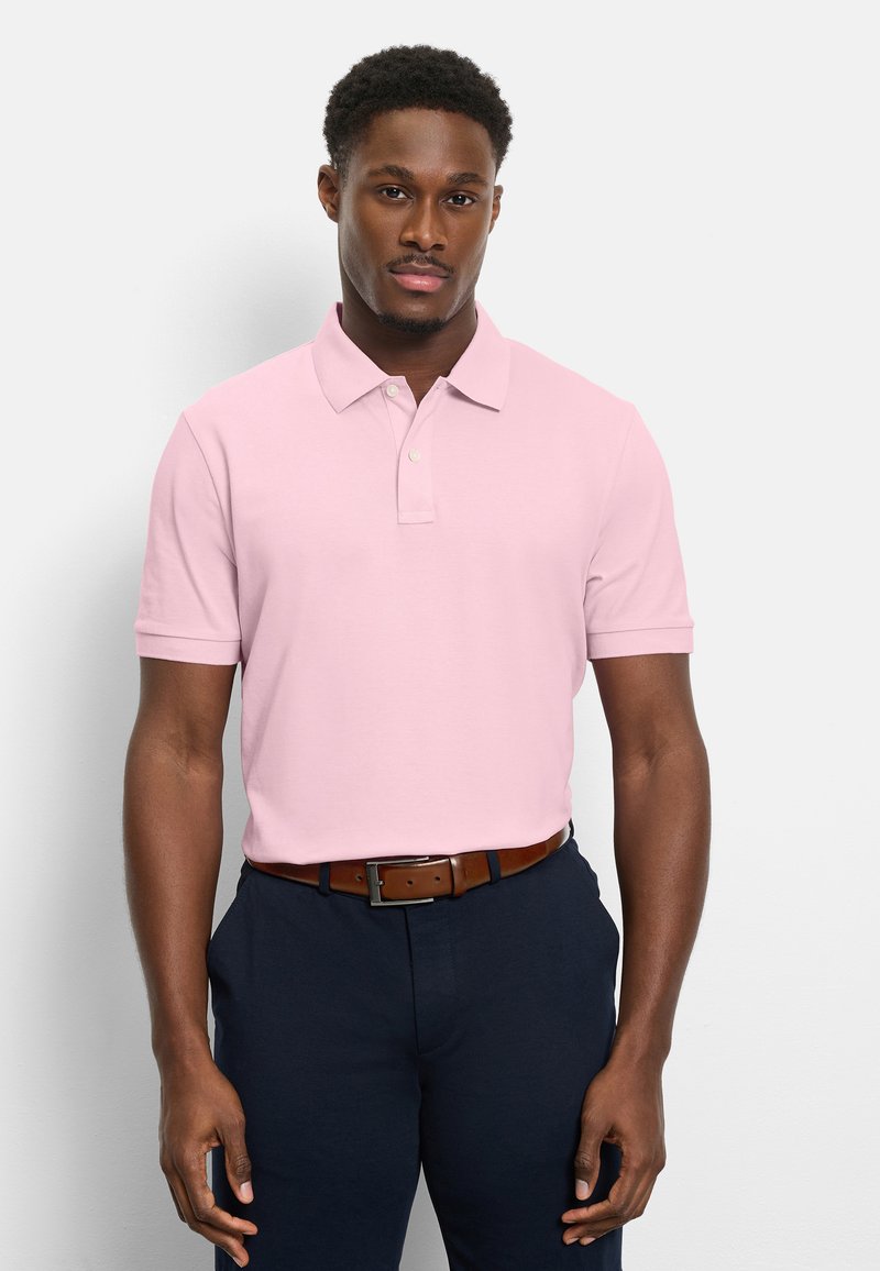 Homme portant un polo à manches courtes rose clair, rentré dans un pantalon foncé avec une ceinture marron, debout devant un fond blanc uni.