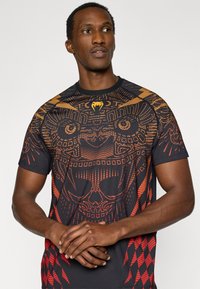 Μαύρο κοντομάνικο μπλουζάκι με περίπλοκα κόκκινα και πορτοκαλί γραφικά μοτίβα, στρογγυλή λαιμόκοψη και υφή που θυμίζει φυλετική τέχνη.