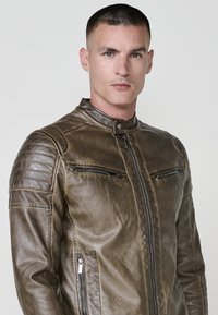 Uomo con capelli corti che indossa una giacca di pelle marrone con tasche con zip, in piedi contro uno sfondo chiaro e semplice, che guarda la macchina fotografica.