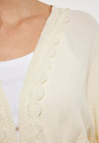 Licht beige cardigan met een geschulpte kantafwerking langs de open voorkant, gelaagd over een witte tanktop met een zachte textuur.