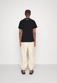 Czarny t-shirt z krótkim rękawem z zieloną metką na plecach, połączony z kremowymi spodniami z elastycznymi mankietami i czarnymi butami typu slip-on.