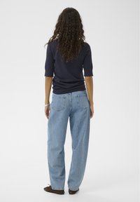 Marineblaues, geripptes Strickoberteil mit kurzen Ärmeln, kombiniert mit locker sitzenden, hellblauen Denim-Jeans. Modell von hinten gesehen, stehend vor einem eintönigen Hintergrund.