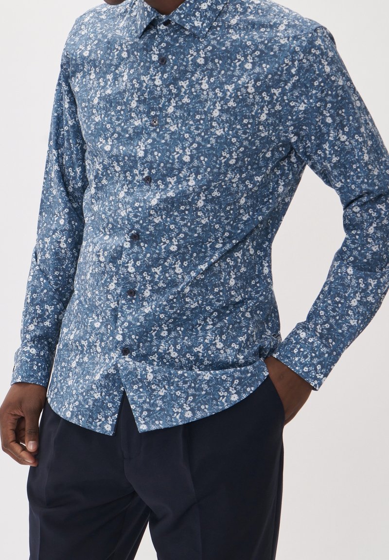 Homme portant une chemise bleue à manches longues fleurie boutonnée et un pantalon foncé, une main dans la poche sur fond uni.