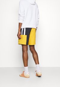Pantaloncini da nuoto gialli e blu marino con una grande tasca laterale, strisce bianche e vestibilità morbida. Abbinati a sneaker beige e calze bianche.