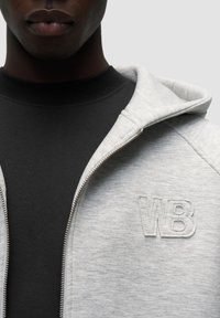 Felpa grigia con zip realizzata in tessuto testurizzato, dotata di un logo ricamato "WB" all'angolo superiore sinistro. Indossata sopra una maglietta nera a girocollo.