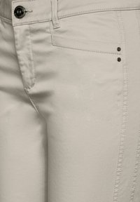 Ljusbeige byxor med en slät yta, som har en framficka och två dekorativa metallnitar. Knäppning och dragkedja vid midjan.