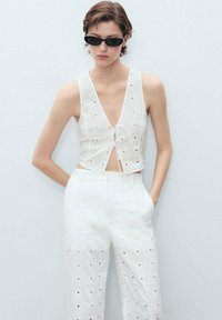 Witte, mouwloze crop top met een diepe V-hals en een strik aan de voorkant. Bijpassende high-waisted broek met een uitsnijdingpatroon.