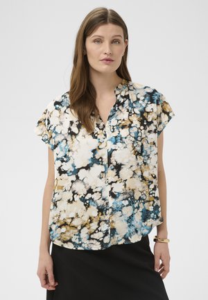 Pruunide pikkade juustega naine kannab lühikeste varrukatega lillelist pluusi sinises, mustas, valges ja beežis, mida täiendavad must seelik ja kuldne käevõru.