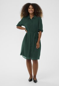 Roheline nööpidega kleit, millel on lipsuvöö, lühikesed rullitud varrukad ja sile, kerge kangas. Seljas mustad terava ninaga jalatsid.