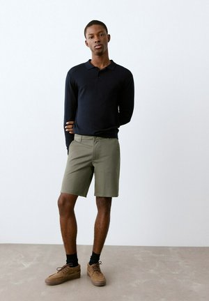 Tânăr stă cu o mână pe spate, purtând un polo cu mâneci lungi de culoare neagră, pantaloni scurți olive, șosete negre și pantofi maro.