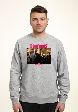 Mężczyzna w szarej bluzie z czerwonym logo "The Sopranos" oraz zdjęciem czterech mężczyzn w garniturach stojących wewnątrz.