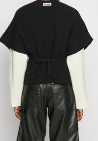 Czarny dzianinowy top z krótkim rękawem, związany w talii. Połączony z białymi, prążkowanymi długimi rękawami i czarnymi skórzanymi spodniami.