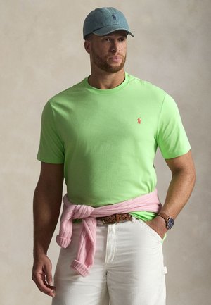 Limezöld pamut póló kis piros logóval, fehér nadrág, és egy rózsaszín pulóver a deréknál megkötve. A kék sapka teszi teljessé az öltözéket.