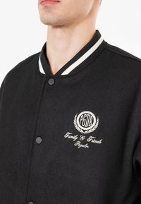Sort varsity-jakke med hvidstribede kraver, med broderet logo og tekst på brystet samt sorte trykknapper. Blød uldlignende tekstur.