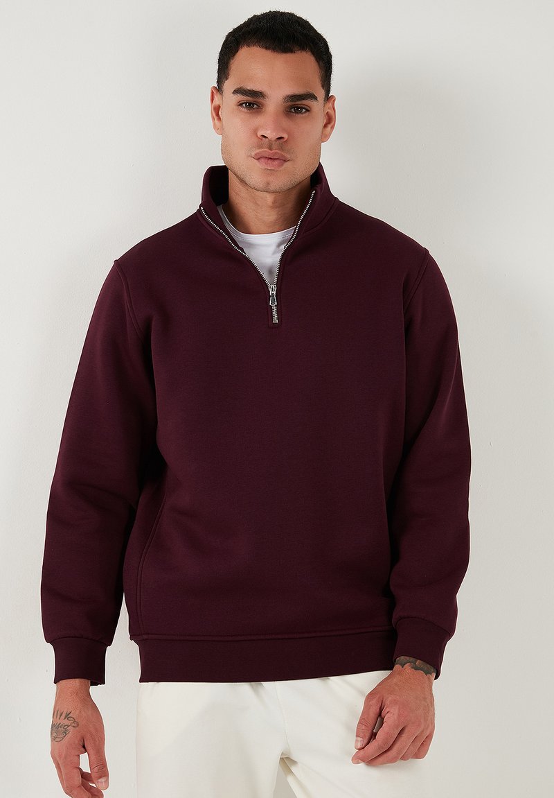 Sudadera burdeos con cremallera y cuello alto, puños acanalados y una textura suave. Presenta un sutil detalle en la costura lateral.
