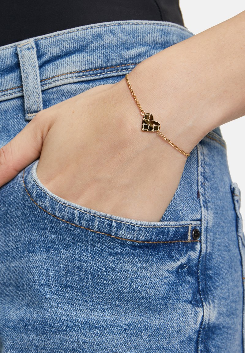 Bracciale in catena d'oro con un ciondolo a forma di cuore nero con motivo a scacchi, indossato su una mano appoggiata in una tasca di denim.