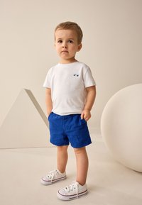 Camiseta blanca de algodón con bordado de coche azul, combinada con pantalones cortos rayados en azul. Zapatillas blancas con detalles en rojo completan el conjunto.