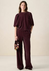 Haut en velours bordeaux avec manches bouffantes et col à volants, assorti d'un pantalon coordonné. Tient un sac à main à motifs, orné d'accents floraux colorés.