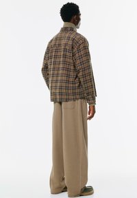 Brązowa koszula w kratę z kołnierzykiem i luźnym krojem, zestawiona z beżowymi spodniami typu wide-leg. Gładka tekstura i kontrastowe detale przyszycia.