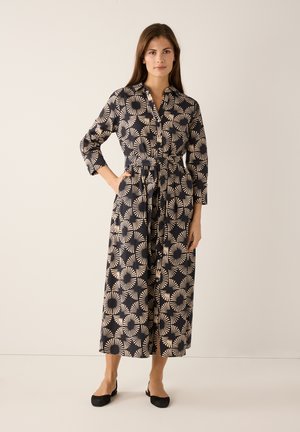 Lange jurk met knopen in marineblauw met een crème cirkelpatroon, met lange mouwen, een strikceintuur en twee zijzakken. Contrasterende zwarte platte schoenen.