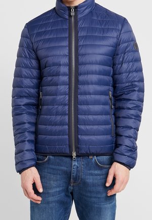 Homme portant une doudoune bleue matelassée avec une fermeture éclair à l'avant et des poches latérales zippées, associée à un jean bleu.