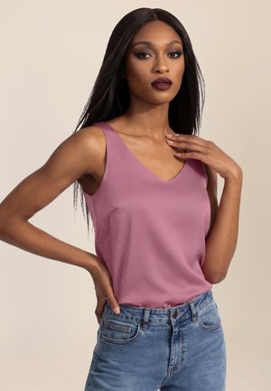 Frau mit langen, glatten Haaren, die ein mauvefarbenes ärmelloses Oberteil und blaue Jeans trägt, posiert mit einer Hand in der Hüfte und der anderen in der Nähe des Schlüsselbeins.