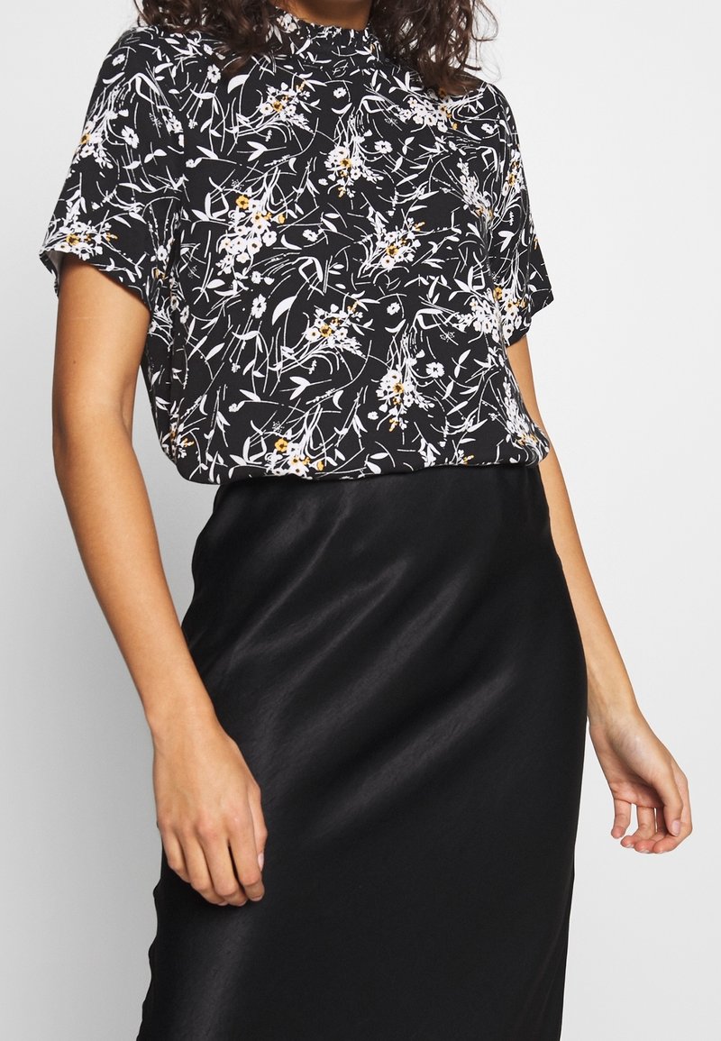 Blouse noire à motifs floraux avec manches courtes, col montant, et motifs floraux blancs et jaunes ; associée à une jupe en satin noire.