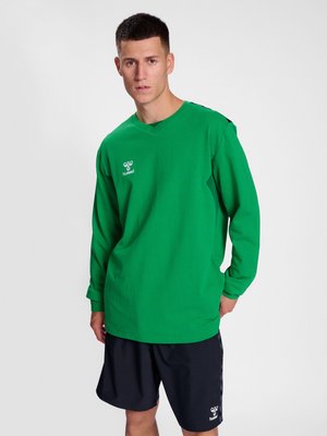 Junger Mann, der ein grünes Langarmshirt und schwarze Shorts trägt, steht vor einem einfarbigen hellen Hintergrund.