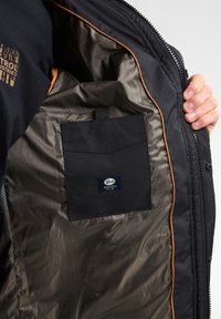 Zwarte jas met een gladde, glanzende bruine voering. Voorzien van een oranje rand, een zwarte pocket en een merketiket.