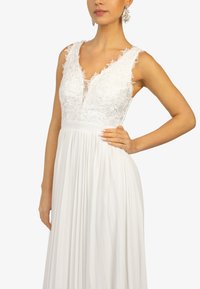 Robe blanche avec un corsage en dentelle au design floral, décolleté plongeant et jupe plissée. Accompagnée de boucles d'oreilles pendantes en argent.