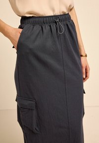 Jupe gris foncé longueur cheville avec taille élastique et cordon de serrage ajustable. Comprend des poches latérales et un tissu lisse et texturé.