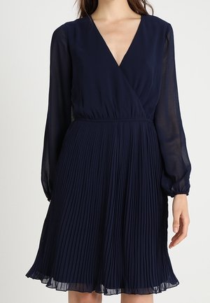 Marineblauwe plisséjurk met lange, doorschijnende mouwen en een diepe V-hals. Vloeiende silhouet en zachte textuur, eindigend boven de knie.