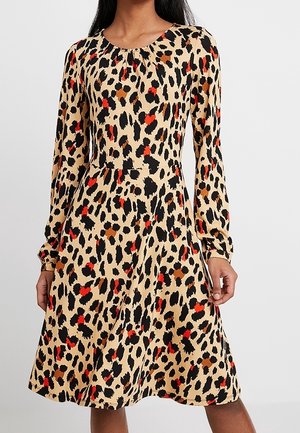 Beige jurk met lange mouwen, voorzien van zwarte, bruine en rode luipaardvlekken, een getailleerde taille en een uitlopende rok, gedragen door een staand persoon.