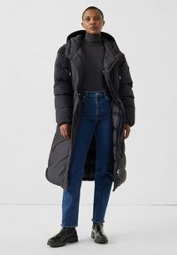 Schwarzer gepolsterter langer Mantel mit Kapuze, kombiniert mit einem taillierten grauen Rollkragenpullover, blauen Jeans und schwarzen Stiefeletten. Glatte Textur und modernes Design.