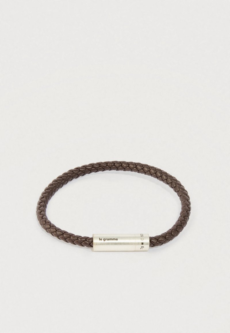 Pīta tumši brūna ādas aproce ar cilindrisku sudraba sprādzi, uz kuras ir iegravēts "le gramme" un svara dati.