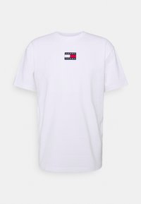 Camiseta de algodón blanca con cuello redondo y mangas cortas. Presenta un pequeño parche de logo colorido "TOMMY JEANS" en el pecho.