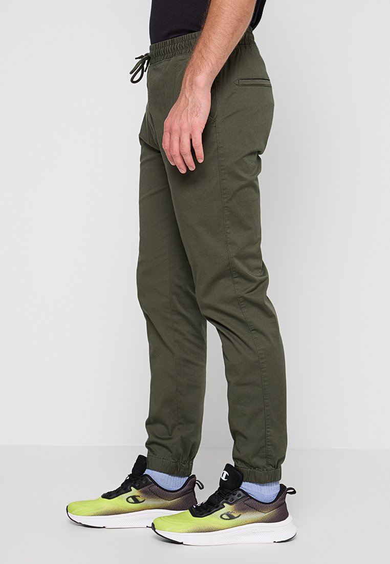 Pantalons de jogging vert olive avec une taille élastique, des poches latérales et des poignets ajustés. Portés avec des chaussures de sport noires et jaunes.