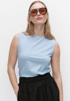 Kobieta o długich blond włosach, nosząca duże brązowe okulary przeciwsłoneczne, jasnoniebieski bezrękawnik i czarną spódnicę z wysokim stanem, stojąca na białym tle.