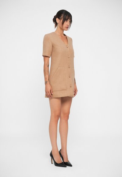 Abito a maniche corte di colore beige con scollatura a V, bottoni sulla parte anteriore, tasche e una vestibilità strutturata sopra le cosce. Indossato con tacchi neri.