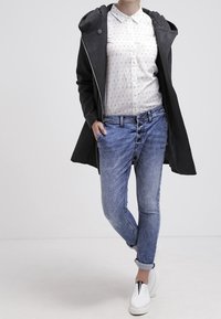 Veste à capuche noire par-dessus une chemise blanche à boutons avec des détails à motifs, jeans en denim bleu et chaussures blanches avec accents noirs.