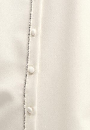 Balta bluzelė, pasižyminti blizgiu paviršiumi, žėrinčiu apvadu mygtukų eigoje ir apvaliais, audiniu aptrauktais mygtukais.