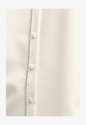 Balta bluzelė, pasižyminti blizgiu paviršiumi, žėrinčiu apvadu mygtukų eigoje ir apvaliais, audiniu aptrauktais mygtukais.