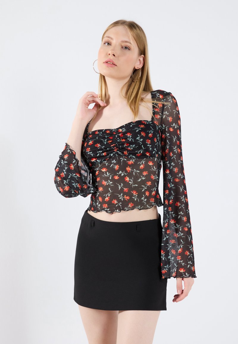Black floral blouse with long, sheer sleeves and ruched front, featuring red and green patterns. Paired with a form-fitting black mini skirt.